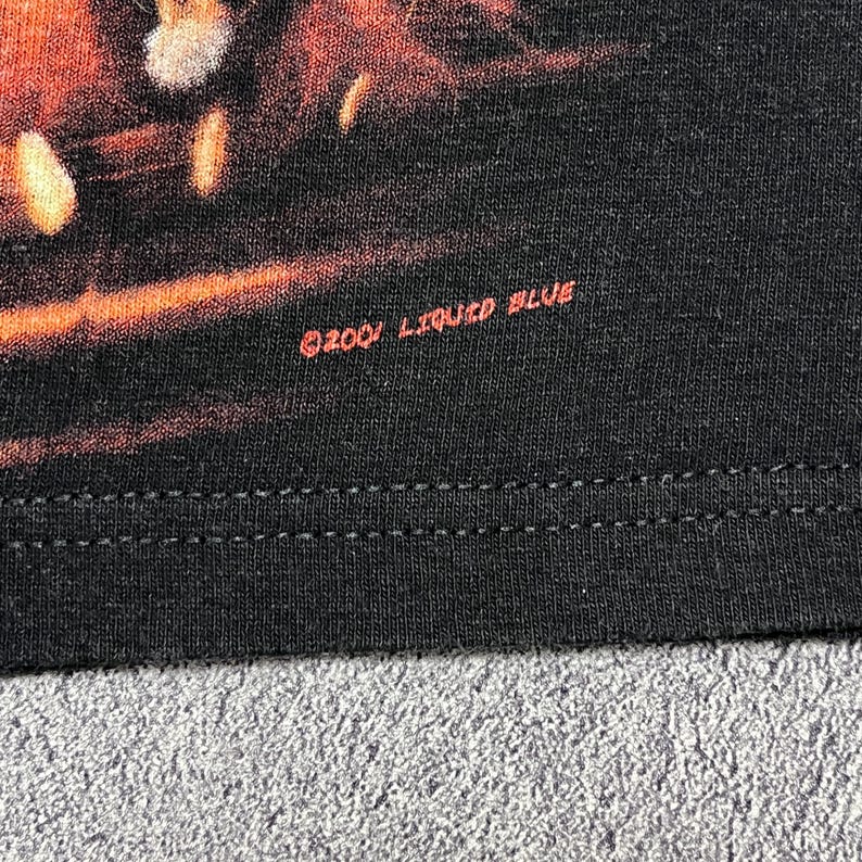 May include: Close-up of a black t-shirt with a graphic design. The text "&copy;2001 LIQUID BLUE" is printed in red on the lower right side of the shirt. The shirt has a double-stitched hem.