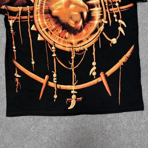 May include: Black t-shirt featuring a large, detailed orange and brown graphic. The graphic includes a circular design with a lion's face and intricate, tribal-inspired elements. The design has a dreamcatcher aesthetic with hanging ornaments.