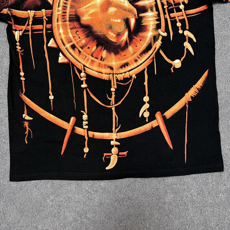 May include: Black t-shirt featuring a large, detailed orange and brown graphic. The graphic includes a circular design with a lion's face and intricate, tribal-inspired elements. The design has a dreamcatcher aesthetic with hanging ornaments.