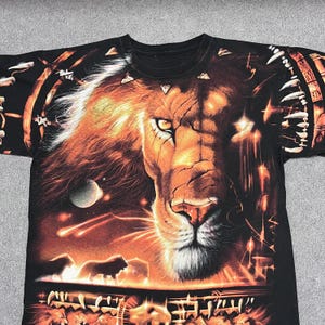 May include: Black t-shirt featuring a detailed, vibrant graphic of a lion's face with a fiery mane. The design includes celestial elements and a lower section with additional lion figures. The shirt has a crew neck.