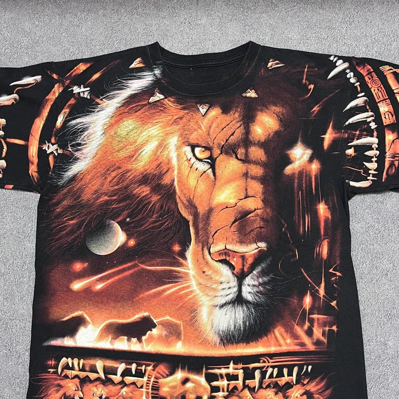May include: Black t-shirt featuring a detailed, vibrant graphic of a lion's face with a fiery mane. The design includes celestial elements and a lower section with additional lion figures. The shirt has a crew neck.
