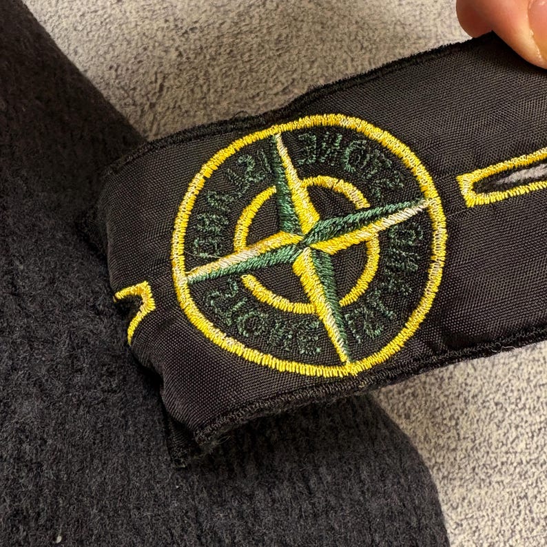 Op de afbeelding: Zwart stoffen embleem met een cirkelvormig embleem met een geel en groen kompasroosontwerp. Het embleem is omgeven door een gele rand met tekst. De patch bedekt gedeeltelijk een donkergrijze stof.