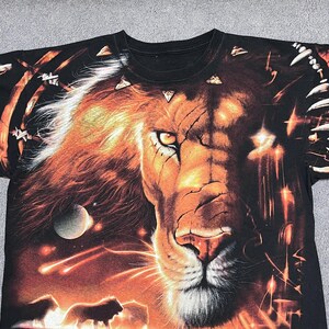 May include: Black t-shirt featuring a detailed graphic of a lion's face with a fiery mane and a cosmic background. The design includes planets, shooting stars, and silhouettes of lions. The shirt is a crew neck.
