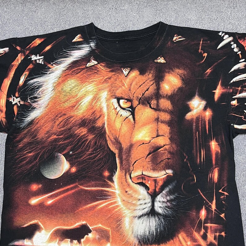 May include: Black t-shirt featuring a detailed graphic of a lion's face with a fiery mane and a cosmic background. The design includes planets, shooting stars, and silhouettes of lions. The shirt is a crew neck.