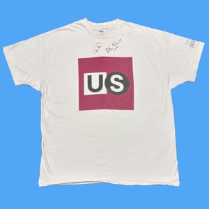 May include: White t-shirt with a maroon square design featuring the letters "US" in white and black. The shirt has handwritten text that says "hello" and "Peter Schmidt".
