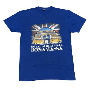 Puede incluir: Camiseta azul real con un gráfico del Royal Albert Hall y una bandera de la Union Jack. El texto "ROYAL ALBERT HALL BONAMASSA" está impreso en blanco debajo. Una camiseta de concierto clásica.