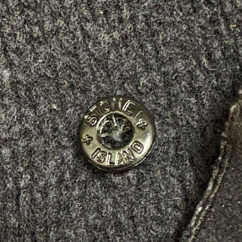 Op de afbeelding: Close-up van een donkergrijze knoop met de woorden "STONE ISLAND" in reli&euml;f langs de rand. De knoop is cirkelvormig en heeft een metallic glans, tegen een achtergrond van een grijze stof.