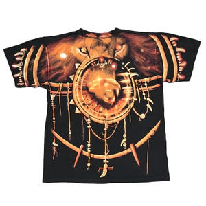May include: Black t-shirt featuring a lion's face with glowing eyes and a circular design. The design includes a lion's head within a circle, with hanging decorative elements. The shirt has a crew neck and short sleeves.