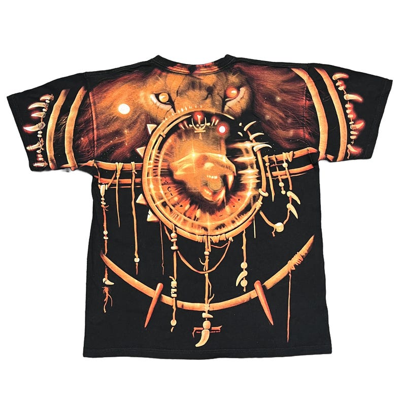 May include: Black t-shirt featuring a lion's face with glowing eyes and a circular design. The design includes a lion's head within a circle, with hanging decorative elements. The shirt has a crew neck and short sleeves.