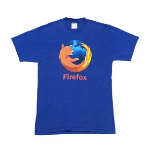May include: Royal blue t-shirt featuring the Firefox logo. The logo depicts a stylized orange fox encircling a blue and white globe, with the word "Firefox" in red below. The shirt is made of a soft material.