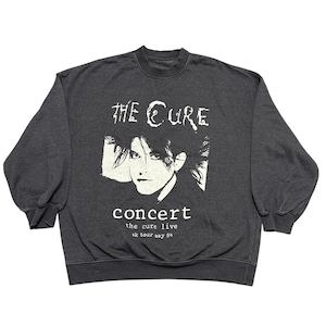 Op de afbeelding: Donkergrijze sweatshirt met een witte afbeelding van een gezicht en de bandnaam "The Cure" boven het woord "concert". Onder het woord "concert" staat de tekst "the cure live 84 tour say". De sweatshirt heeft lange mouwen.