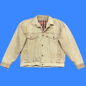 May include: A tan denim jacket with a red plaid lining peeking through the collar. The jacket has two chest pockets, two side pockets, and a button-up front. A small red tag is visible on the left chest pocket.
