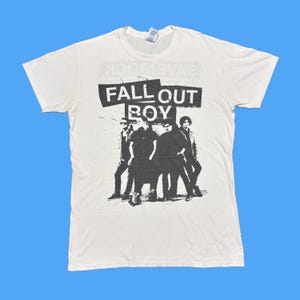 Könnte beinhalten: Cremefarbenes T-Shirt mit dem Bandnamen "FALL OUT BOY" in einem schwarzen Rechteck über einem Schwarz-Weiß-Bild der Bandmitglieder. Das T-Shirt hat einen Rundhalsausschnitt.