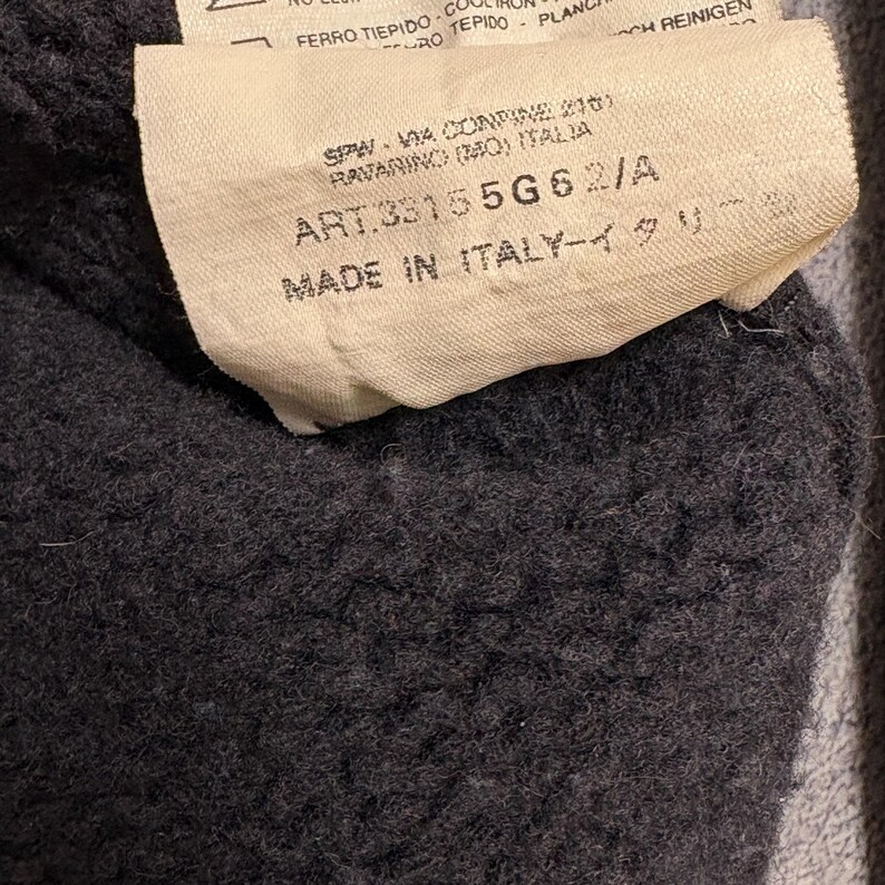 Op de afbeelding: Close-up van een donkergrijze stof met een beige label eraan. Het label heeft zwarte tekst, waaronder "MADE IN ITALY". De stof heeft een getextureerde, fuzzy uitstraling.