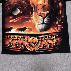 May include: Black t-shirt featuring a detailed graphic of a lion's face and other lions in a fiery, orange-toned design. The artwork includes a celestial background with a moon and shooting stars, creating a dramatic effect.