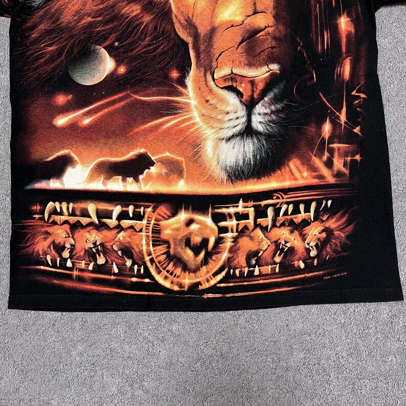 May include: Black t-shirt featuring a detailed graphic of a lion's face and other lions in a fiery, orange-toned design. The artwork includes a celestial background with a moon and shooting stars, creating a dramatic effect.