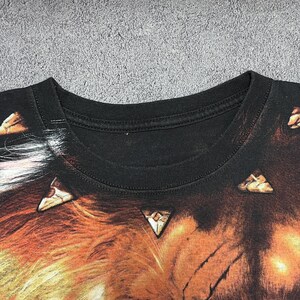 May include: Black crew neck t-shirt featuring a detailed graphic of a lion's face with orange, brown, and white fur. The design includes triangular accents. The shirt's collar is black, and the fabric appears to be cotton or a similar material.