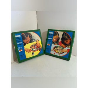 Può includere: Due scatole di puzzle in legno vintage con bordi verdi e blu. Ogni scatola presenta un bambino che lavora al puzzle. Le scatole hanno il testo "Wooden Puzzle" e "uten puzzle". I puzzle sono progettati per bambini dai 3 anni in su.
