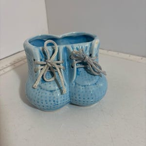 Könnte beinhalten: Hellblauer Keramik-Babyschuh-Pflanztopf mit weißen Schnürsenkeln. Der Pflanztopf hat eine strukturierte Oberfläche und ist als Paar Babyschuhe gestaltet. Der Pflanztopf ist ein dekorativer Artikel.