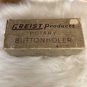 May include: A vintage rectangular box with the text "GREIST Products ROTARY BUTTONHOLER" in brown lettering. The box has a textured, woven appearance in shades of tan and brown, with some wear and tear visible on the edges.