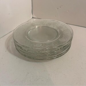 Puede incluir: Una pila de platillos de vidrio transparente con un diseño floral grabado alrededor del borde. Los platillos están apilados uno encima del otro, con el platillo superior enfocado. El fondo es una superficie blanca lisa.