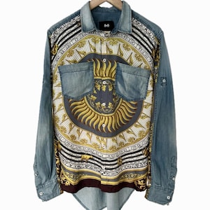 May include: A long-sleeve denim shirt featuring a central panel with a gold sun graphic, zodiac symbols, and calendar dates. The shirt has a button-down front, two chest pockets, a collar, and a D&G label on the inner neck. The sleeves and back are blue denim.