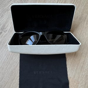 Może przedstawiać: Czarne okulary przeciwsłoneczne z białym etui i czarną ściereczką do czyszczenia. Okulary przeciwsłoneczne znajdują się w białym etui z nazwą marki "VERSACE" wydrukowaną wewnątrz. Czarna ściereczka do czyszczenia z nazwą marki "VERSACE" znajduje się poniżej.