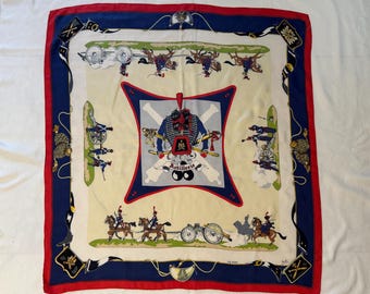 May include: A square silk scarf with a military theme. The scarf features a cream-colored center with illustrations of soldiers, cannons, and horses. The border is blue and red, with decorative elements in the corners. The word "Artillerie" is visible.