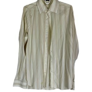 Könnte beinhalten: Cremefarbenes Button-Down-Hemd mit vertikalen Streifen in Braun und Marineblau. Das langärmlige Hemd hängt an einem weißen Kleiderbügel. Das Hemd hat einen klassischen Kragen und geknöpfte Manschetten.