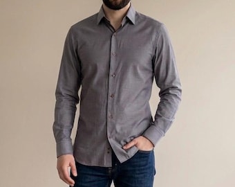 Könnte beinhalten: Ein graues Hemd mit Knöpfen, langen Ärmeln und Kragen. Das Hemd wird mit dunkelblauen Jeans kombiniert. Die Person steht vor einem neutralen Hintergrund.