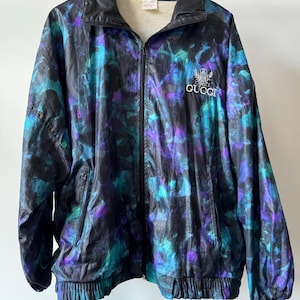 Puede incluir: Chaqueta vintage con cremallera y un vibrante estampado abstracto en verde azulado, morado y negro. La chaqueta presenta cuello y puños negros, un logotipo bordado blanco y un dobladillo acanalado. La chaqueta está colgada en una percha de madera.