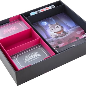 Op de afbeelding: Een zwarte speldoos met roze binnencompartimenten. De doos bevat kaarten met de tekst "CASTING SHADOWS", dobbelstenen en een spelbord met een cartoonwolf. De speldoos is ontworpen voor het opbergen en organiseren van bordspellen.