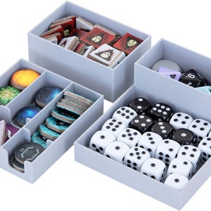 Pode incluir: Quatro caixas de armazenamento de plástico cinza com divisórias, cheias de várias peças de jogo, incluindo dados, fichas e cartas. As caixas são projetadas para organizar e armazenar os componentes do jogo.