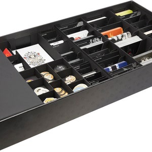 Può includere: Scatola di stoccaggio nera con scomparti per organizzare i pezzi del gioco. La scatola ha un coperchio ed è piena di vari componenti del gioco, tra cui carte, gettoni e dadi.