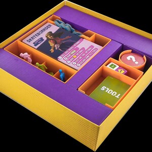Op de afbeelding: Paarse en oranje game organizer insert met vakken voor kaarten, dobbelstenen en kleine figuurtjes. De insert is gelabeld "Skateboarder" en "Tools".