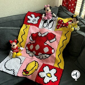 Peut inclure: Une couverture au crochet sur le th&egrave;me de Mickey Mouse. La couverture est compos&eacute;e de carr&eacute;s color&eacute;s avec des images des gants de Mickey Mouse, une robe, une fleur et un chapeau. Il y a trois peluches Mickey Mouse sur la couverture.