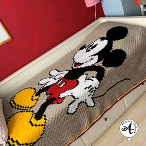 Könnte beinhalten: Eine gehäkelte Decke mit Mickey Mouse in einer klassischen Pose. Die Decke ist beige mit schwarzen, weißen, roten und gelben Akzenten. Die Decke hat einen gehäkelten Rand mit roten, weißen und gelben Streifen.