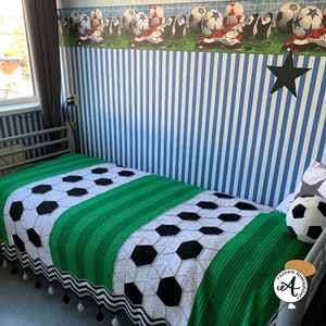 Może przedstawiać: Zielono-biały koc wykonany na szydełku z wzorem piłki nożnej. Koc ma czarno-białe paski wokół krawędzi i czarne pompony.