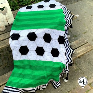 May include: A green, white, and black crocheted blanket with a soccer ball design. The blanket has a black and white zigzag trim and white pom-poms.