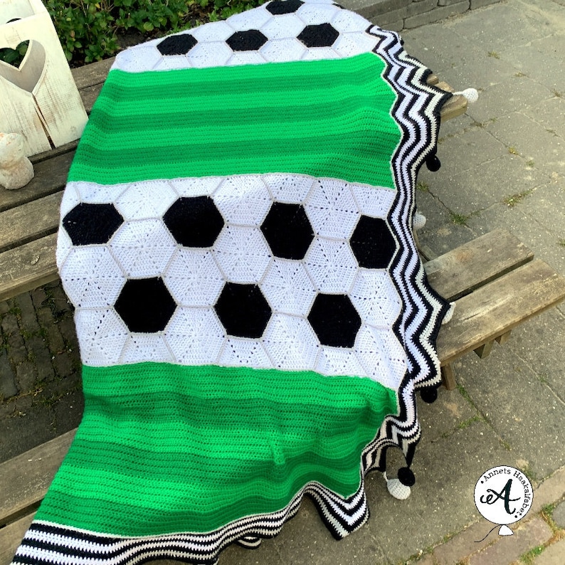 May include: A green, white, and black crocheted blanket with a soccer ball design. The blanket has a black and white zigzag trim and white pom-poms.