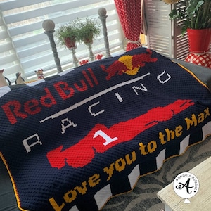 Op de afbeelding: Een blauwe en rode gehaakte deken met de woorden "Red Bull Racing" en "Love you to the Max" in wit. De deken heeft een rood en geel stierenlogo en een rode raceauto met nummer 1.