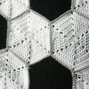 May include: Close-up of a white crocheted soccer ball blanket. The blanket is made of individual hexagonal crochet stitches, with a gray border around each hexagon. The blanket is partially covering a black background.