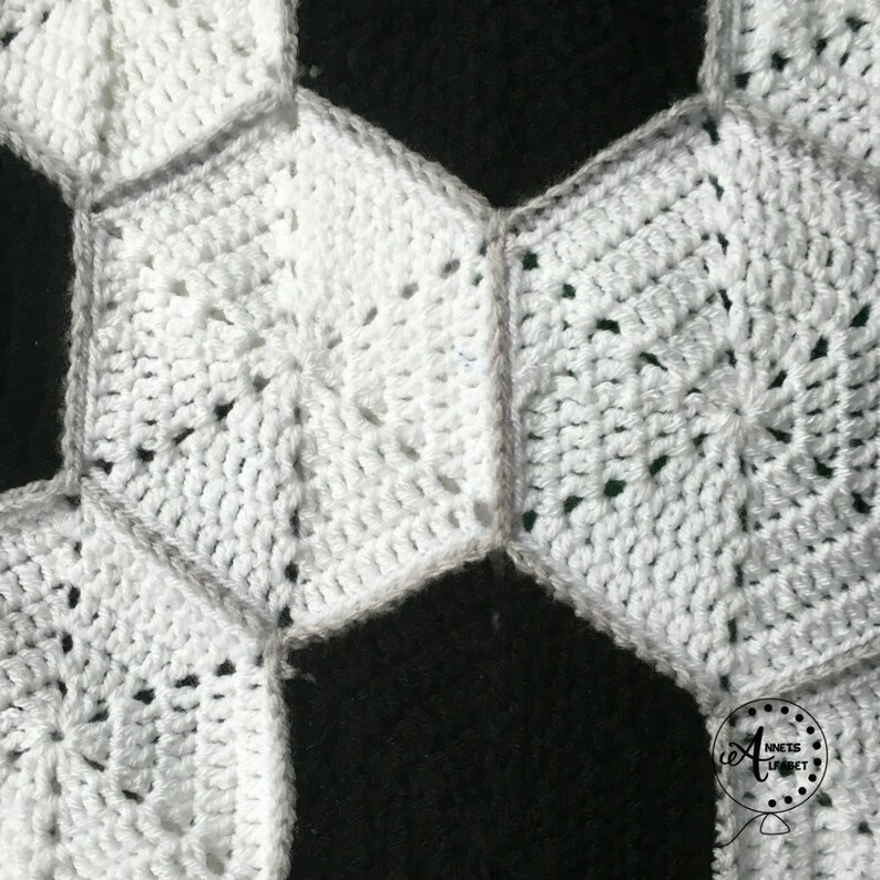 May include: Close-up of a white crocheted soccer ball blanket. The blanket is made of individual hexagonal crochet stitches, with a gray border around each hexagon. The blanket is partially covering a black background.