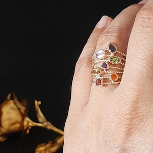 Puede incluir: Un conjunto de anillos de oro con piedras preciosas de colores. Los anillos están apilados en un dedo y presentan una variedad de formas y tamaños de piedras, incluyendo una piedra roja rectangular, una piedra naranja redonda, una piedra púrpura en forma de lágrima, una piedra blanca rectangular, una piedra verde redonda y una piedra púrpura en forma de lágrima.