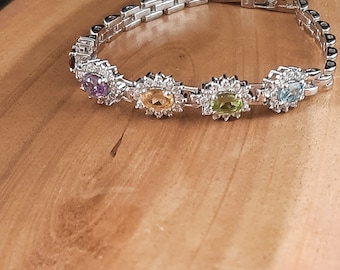 Può includere: Un braccialetto d'argento con un design a catena a maglie e cinque pietre ovali incastonate in un motivo floreale. Le pietre sono viola, gialle, verdi, blu e trasparenti.