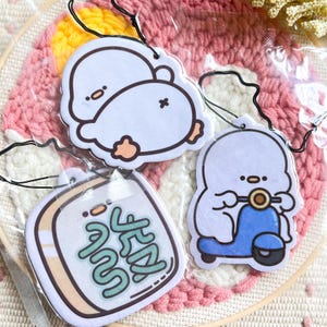 Könnte beinhalten: Drei weiße Auto-Lufterfrischer mit Comicfiguren. Einer zeigt einen Pinguin mit einem Herz auf dem Rücken, ein anderer einen Pinguin, der einen Roller fährt, und der letzte ein Quadrat mit dem Text "Good Vibes" in Grün.