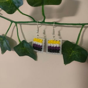 Op de afbeelding: Een paar oorhangers met een geel, wit, paars en zwart kruissteekontwerp. De oorhangers hangen aan zilveren haakjes.