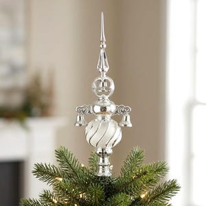 Könnte beinhalten: Ein silberner Weihnachtsbaumspitzen-Aufsatz mit einer hohen, spitzen Finial, einer zentralen weißen Glaskugel mit Spiralmuster und drei kleinen hängenden Glocken. Die Struktur ist metallisch und reflektierend und sitzt auf grünen Tannenzweigen.
