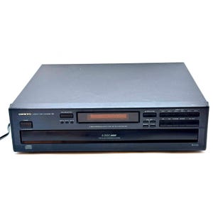 May include: A black Onkyo DX-C100 compact disc changer featuring a 6-disc capacity, a digital display, and multiple control buttons on the front panel. The unit includes labels for disc selection, playback controls, and power status.