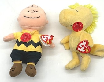 Może przedstawiać: Dwie pluszowe zabawki przedstawiające Charliego Browna i Woodstocka. Charlie Brown ma na sobie żółtą koszulkę w czarny zygzak, czarne szorty i brązowe buty. Woodstock to żółty ptak z czubkiem na głowie. Obie zabawki mają czerwone metki Ty.